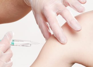 Ktoś w białych, jednorazowych rękawiczkach podaje szczepionkę w ramię innej osoby.
