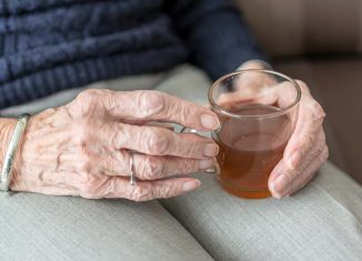 Na zdjęciu dłonie starczej kobiety, trzymającej kubek z herbatą.